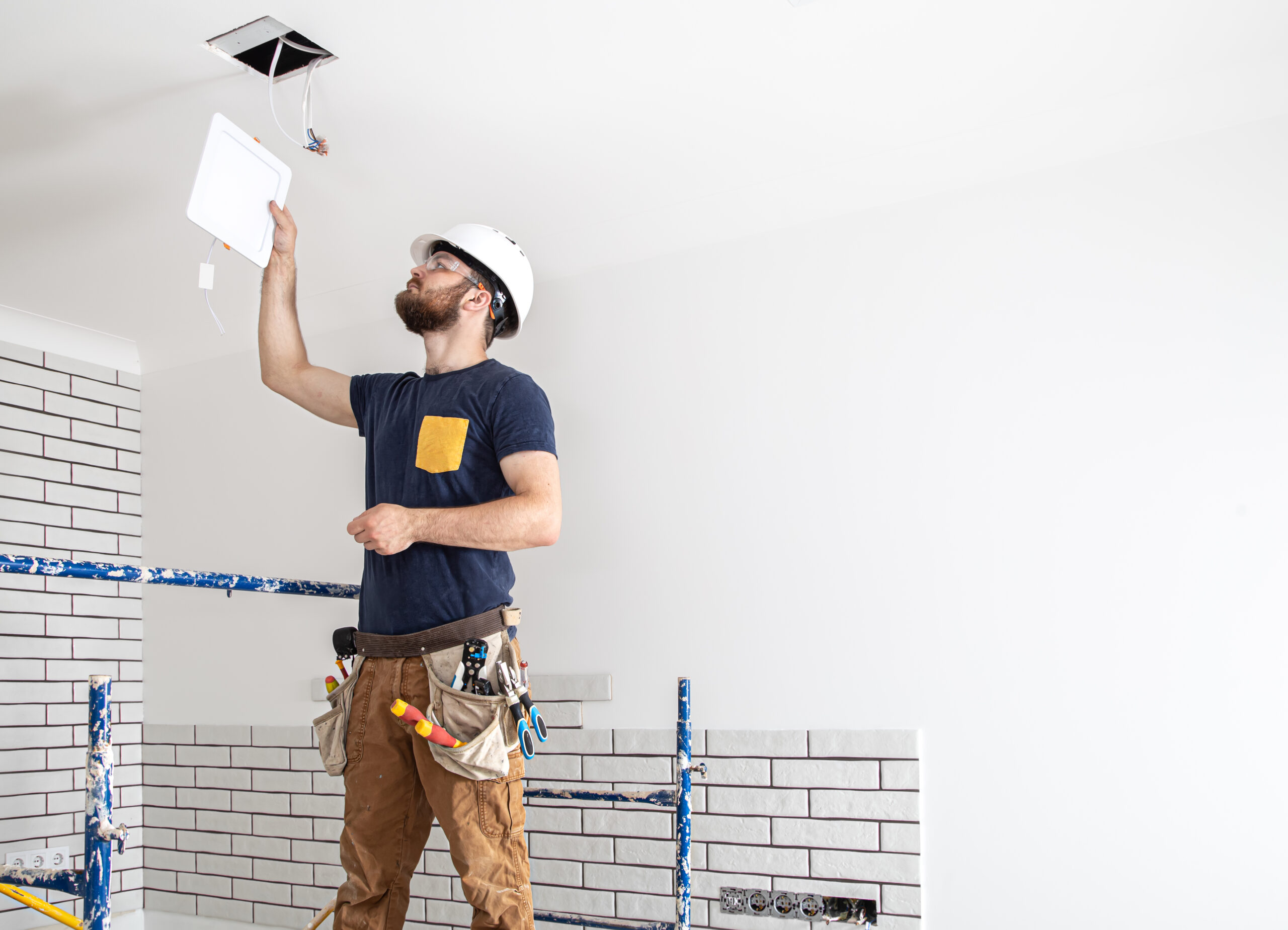 Electrician Builder with beard worker in a white helmet at work, installation of lamps at height. Professional in overalls with a drill on the background of the repair site.